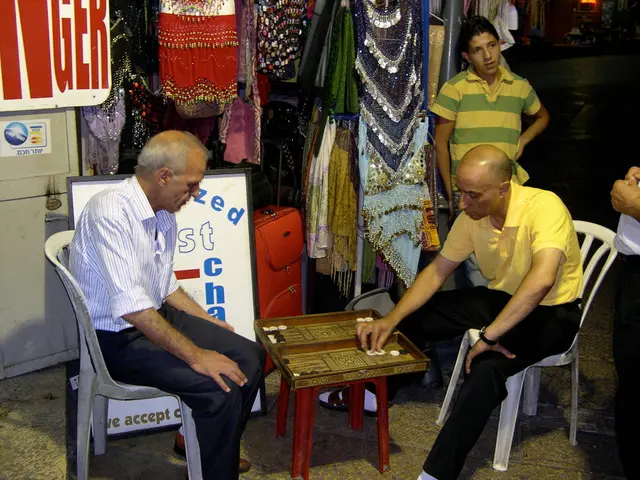 In this image we can see two men are sitting on the chair and playing a board game which is on the...